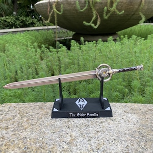 Pu&ograve; includere: Una spada decorativa del franchise "The Elder Scrolls", esposta su un supporto nero. La spada presenta una lama color bronzo, un'elsa argentata dettagliata e un'impugnatura nera. Il supporto presenta il logo del gioco e il testo.