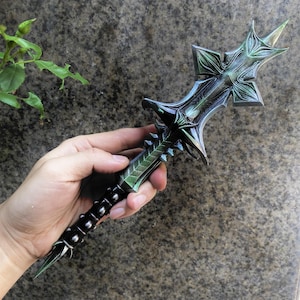 May include: A detailed, fantasy-style dagger with a green and black color scheme. The blade has a cross-shaped hilt and a spiked handle. The dagger is held in a hand against a mottled gray background.