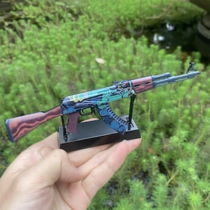 May include: A miniature AK-47 model with a decorative design. The model features a wooden stock and handguard, with a colorful, ornate pattern on the receiver and magazine. The model is displayed on a black stand against a green background.