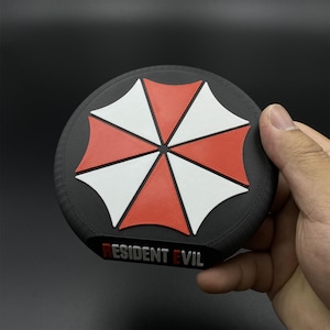 May include: A black, round coaster featuring a red and white umbrella design. The coaster has the text "RESIDENT EVIL" in a rectangular box at the bottom. The coaster is held by a hand against a black background.
