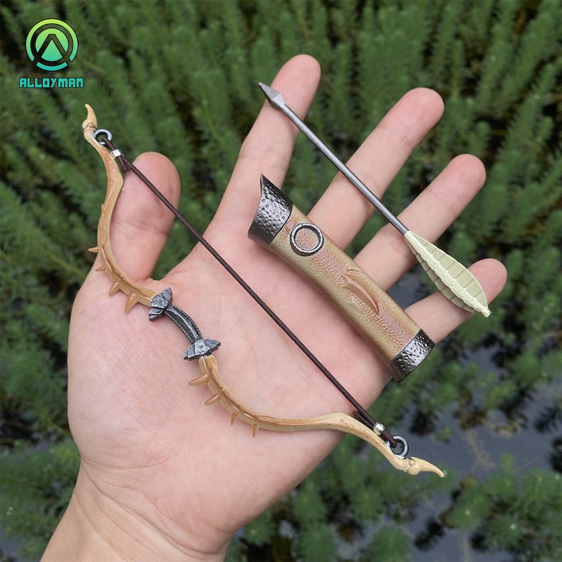 Puede incluir: Un juego de arco y flecha en miniatura con un carcaj. El arco es de color marr&oacute;n claro con una cuerda oscura y detalles decorativos. El carcaj es de color beige con una parte superior e inferior de metal oscuro. Tambi&eacute;n se incluye una flecha con punta plateada y plumas verdes.