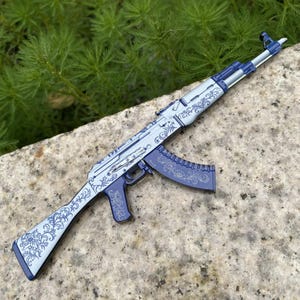 May include: A decorative AK-47 style rifle in shades of blue and silver, adorned with intricate floral patterns. The rifle is resting on a stone surface, with green foliage in the background.