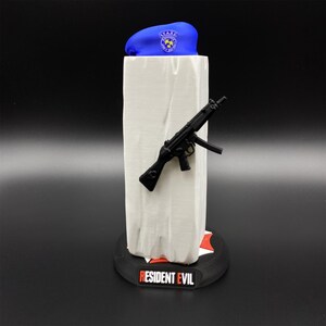 May include: A blue beret with a badge, a black toy gun, and a white rectangular object are displayed on a black base with the text "RESIDENT EVIL". The beret has a badge with stars.