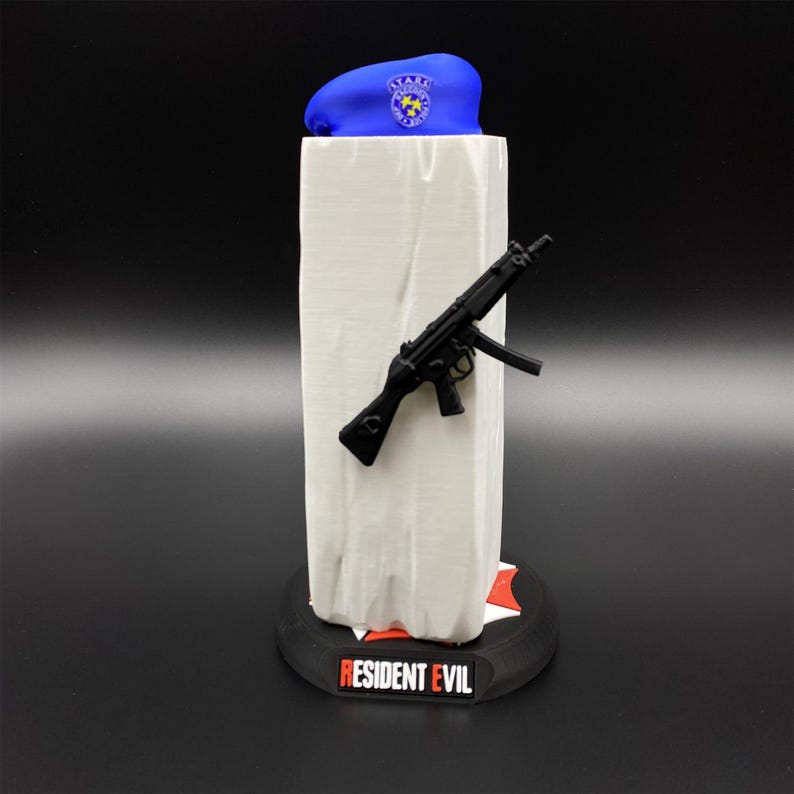 May include: A blue beret with a badge, a black toy gun, and a white rectangular object are displayed on a black base with the text "RESIDENT EVIL". The beret has a badge with stars.