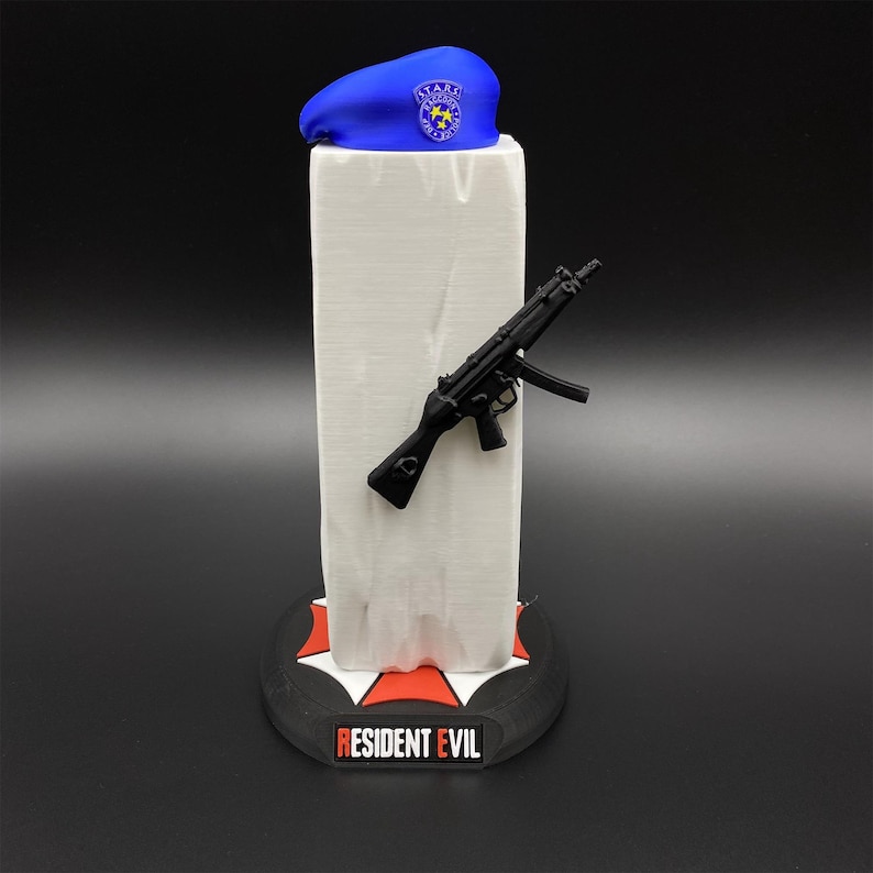 May include: A blue beret with a STARS emblem sits atop a white rectangular structure. A black toy gun is attached to the structure. The base is black with a red and white emblem and the words "RESIDENT EVIL".