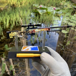 May include: A miniature sniper rifle model featuring a yellow and orange dragon-themed pattern on the stock and receiver, equipped with a black scope and barrel. The model is mounted on a black display stand with a gold bullet and a blue logo.