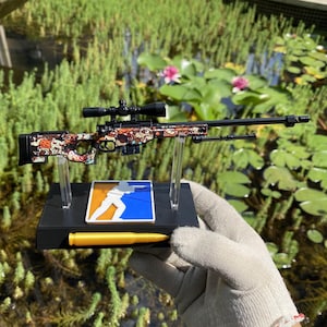 Könnte beinhalten: Ein Miniatur-Scharfschützengewehr-Modell mit einem bunten Cartoon-Figuren-Muster auf Schaft und Lauf. Das Gewehr ist auf einem schwarzen rechteckigen Ständer mit einem blau-orangen Logo und einer gelben Patronennachbildung montiert.
