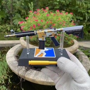 May include: A miniature rifle model featuring a gold and silver camouflage pattern, mounted on a black display stand. The stand includes a blue and orange square emblem with a white silhouette and a horizontal yellow cylindrical accent piece.