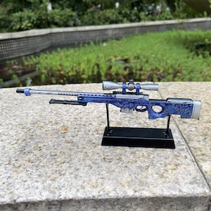 May include: A miniature sniper rifle with a blue and silver finish, featuring detailed engraved patterns. The scope is silver, and the rifle is mounted on a black stand. The background is blurred, with green foliage.