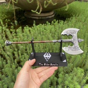 May include: A silver and bronze axe with intricate designs, displayed on a black stand. The stand features the text "The Elder Scrolls" and a logo. The axe has a detailed blade and a decorative handle.