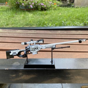 Op de afbeelding: Een wit en zwart speelgoed sluipschuttergeweer met een vizier. Het geweer heeft zwart-witte grafische ontwerpen en is gemonteerd op een zwarte standaard. De achtergrond is een houten bank.