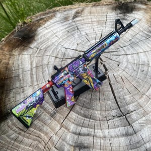 以下が含まれることがあります： 漫画のキャラクターとグラフィティ風のアートが特徴の、鮮やかなデザインのカラフルなおもちゃのライフル。ライフルは主に紫、赤、黄色で、黒のアクセントがあります。黒いスタンドに展示されています。