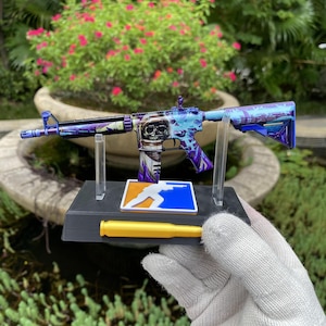 Könnte beinhalten: Ein Miniatur-Gewehrmodell mit violett-blauem kosmischem Muster und einer Totenkopf-Grafik auf dem Gehäuse. Das Modell ist auf einem schwarzen Ständer montiert, der einen gelben, patronenförmigen Akzent und ein quadratisches Logo zeigt.