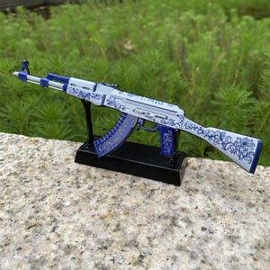 May include: A decorative AK-47 rifle replica. The rifle features a white and blue floral pattern. The replica is mounted on a black stand. The background is blurred green foliage.