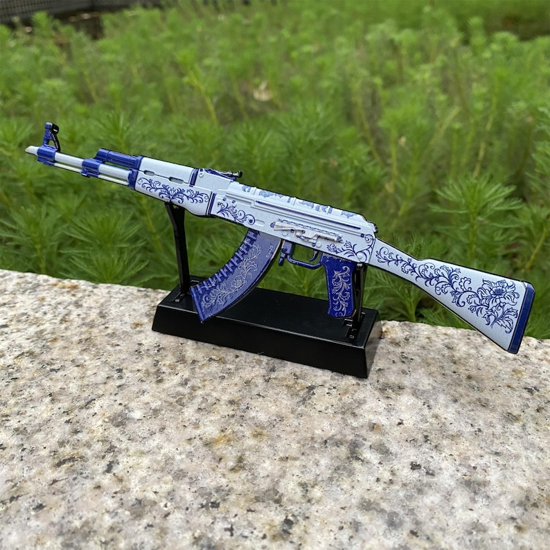 May include: A decorative AK-47 rifle replica. The rifle features a white and blue floral pattern. The replica is mounted on a black stand. The background is blurred green foliage.