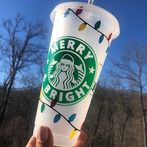 May include: White reusable cup with a green Starbucks logo and the text "Merry Bright" surrounded by a string of colorful Christmas lights.