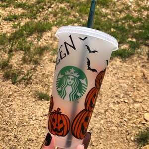 May include: A white reusable cup with a black and green Starbucks logo and a Halloween theme. The cup is decorated with orange pumpkins and black bats. The text "EEN" is visible on the cup.