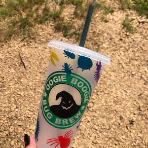 May include: A clear plastic reusable cup with a green, white and black design featuring the text "Oogie Boogie Bug Brew" and a silhouette of Oogie Boogie from the film "The Nightmare Before Christmas". The cup has a black lid and a blue and green straw.