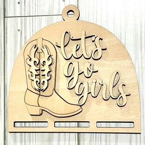 May include: Wooden sign with a cutout of a cowboy boot and the words "let's go girls" in cursive. The sign is light brown with a rounded top and a small hole for hanging. The boot has decorative cutouts.