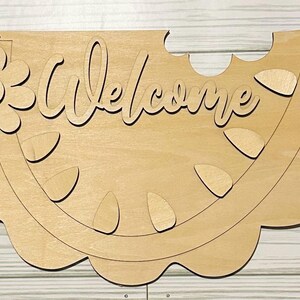 May include: Wooden cutout of a watermelon slice with the word "Welcome" in a cursive font. The watermelon has a flower with a button in the center on the left side.