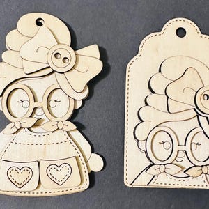 May include: Two wooden cutouts of a cartoon character with glasses, a bow, and a button. One cutout is a full figure with heart-shaped pockets. The other is a tag-shaped cutout of the character's face.