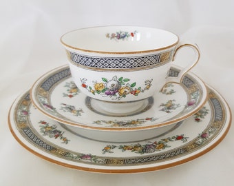 May include: A white teacup and saucer set with a floral pattern and gold trim. The cup and saucer have a blue and gold border with a diamond pattern.