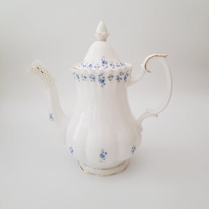 May include: A white porcelain teapot with a floral pattern of blue forget-me-nots. The teapot has a gold trim around the lid and base.