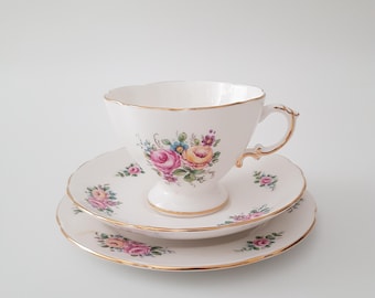 May include: A white teacup and saucer set with a floral design featuring pink, yellow, and blue flowers. The set has gold trim and a scalloped edge. The teacup sits on a matching saucer and a smaller plate.