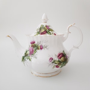 May include: White porcelain teapot with a gold trim and a floral design featuring purple and green thistles.