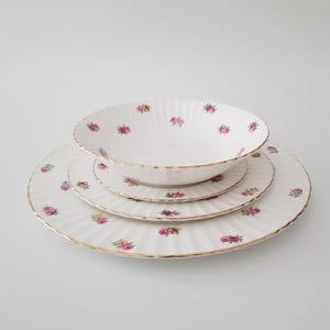 May include: A white porcelain dinner set with a floral design of pink and purple roses. The set includes a large plate, a medium plate, and a bowl, all with gold edging. The plates have a ridged pattern.