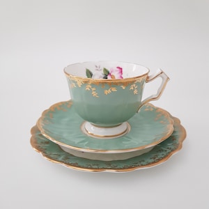 May include: A sage green porcelain teacup and saucer set featuring a scalloped edge and intricate gold leaf detailing along the rims. The interior of the teacup displays a small floral motif with pink and white petals and green leaves.
