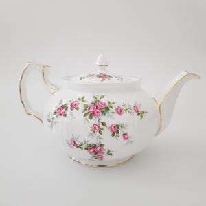 May include: A white porcelain teapot with a lid, decorated with pink rose floral patterns and gold edging. The teapot has a curved spout and handle. A classic design for serving tea, ideal for afternoon tea parties.