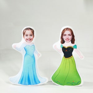 Op de afbeelding: Twee gepersonaliseerde prinsessenkussens met gezichten van kinderen. Het kussen links draagt een blauwe jurk met een wit lijfje en een witte kroon. Het kussen rechts draagt een groene jurk met een zwart lijfje en een rode kroon.