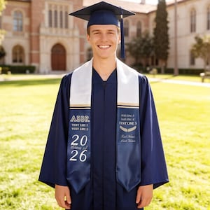 May include: A graduate in a navy blue gown and cap, with a white and navy blue sash. The sash has text that reads "ABBR", "2026", and "Class of 26". The background shows a building and green grass.