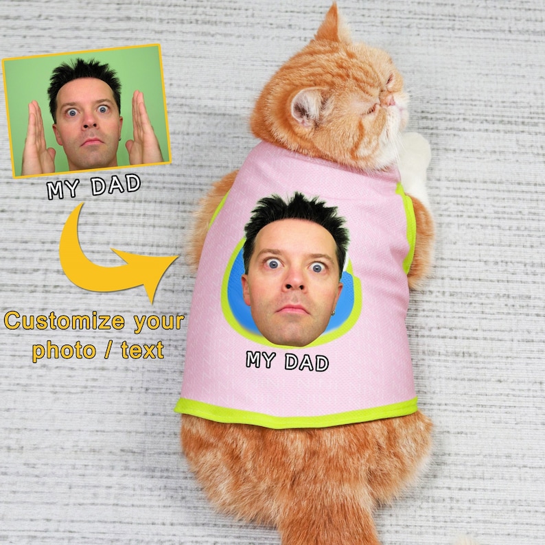 May include: A pink cat vest with a green trim and a blue circle with a man's face in it. The text "MY DAD" is printed on the vest. The vest is on a cat.