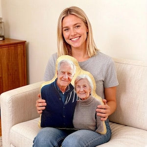 Puede incluir: Un cojín personalizado con una fotografía impresa de una pareja de ancianos. El cojín tiene un borde dorado y la pareja está sonriendo. El cojín lo sostiene una persona que lleva una camiseta gris y vaqueros azules.