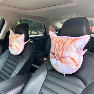 K&ouml;nnte beinhalten: Zwei Kopfst&uuml;tzen f&uuml;r Autos mit einem pinkfarbenen und wei&szlig;en Katzenkopfdesign. Die Katze hat orangefarbenes und wei&szlig;es Fell und geschlossene Augen.