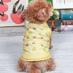 May include: A small brown dog wearing a yellow sleeveless shirt with a cat and duck print. The shirt has the word "NINI" repeated on the bottom.
