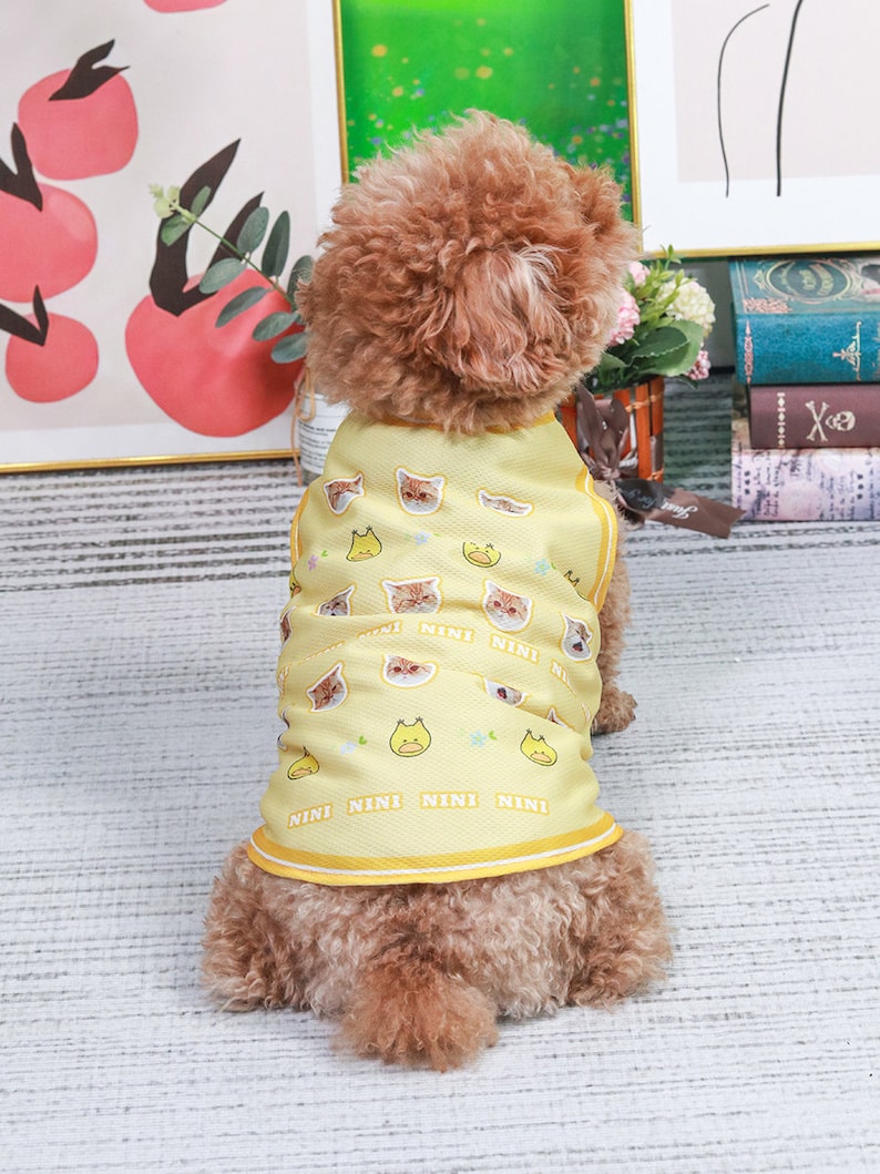 May include: A small brown dog wearing a yellow sleeveless shirt with a cat and duck print. The shirt has the word "NINI" repeated on the bottom.