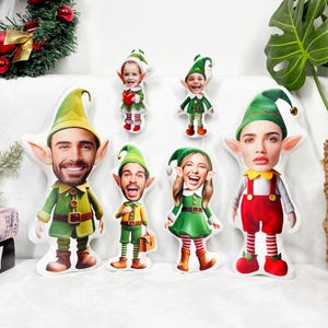 Puede incluir: Una colección de muñecos de duendes de peluche con caras personalizadas. Los muñecos visten trajes verdes y rojos, sombreros de duende y diversas expresiones faciales. Algunos muñecos sostienen objetos pequeños. Fondo con una corona navideña.