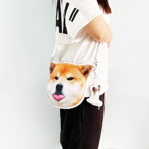 Puede incluir: Bolso bandolera blanco con un diseño realista de la cara de un perro Shiba Inu, con la lengua fuera. El bolso incluye un llavero a juego con forma de perro. El pelaje del perro es una mezcla de marrón, beige y blanco.