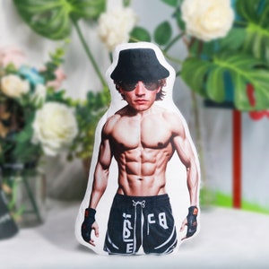 May include: A white pillow featuring a printed image of a muscular man wearing a black hat, sunglasses, and black shorts with white accents. The man has a muscular physique and is posing with his arms outstretched.