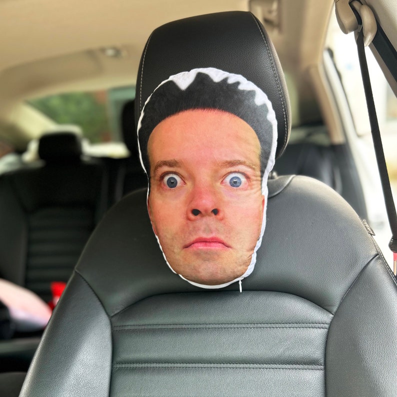 K&ouml;nnte beinhalten: Ein wei&szlig;es Kissen mit einem aufgedruckten Bild eines menschlichen Gesichts. Das Kissen befindet sich in einem Autositz.