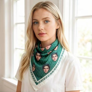 May include: A green and white silk scarf featuring a repeating pattern of faces. The scarf has a diamond pattern in green with a white border and a decorative edge. The scarf is worn around the neck.