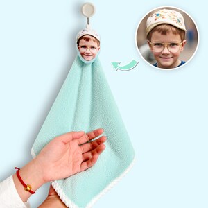Puede incluir: Una toalla de mano azul claro con un borde blanco, colgada de un gancho. La parte superior de la toalla presenta una imagen impresa de un niño con gafas y un sombrero. Una segunda imagen del niño está en un círculo a la derecha.