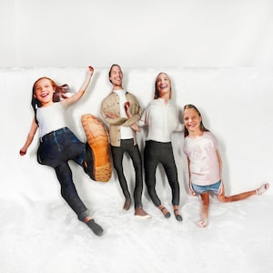 May include: A family of four, represented as soft, plush pillows, arranged on a white surface. The pillows have realistic faces and clothing, with one child kicking a large boot. The other family members are standing and smiling.