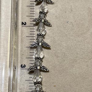 Peut inclure: Un bracelet de perles transparentes avec des accents d'ailes d'ange argentés. Le bracelet mesure environ 12,7 cm de long et est mesuré à côté d'une règle.