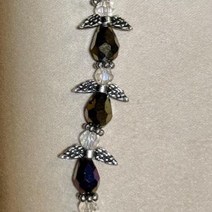 May include: A beaded angel ornament with faceted black and clear beads. Each angel has silver-toned wings. The ornament is strung together with silver-toned wire. The angels are arranged in a vertical line.