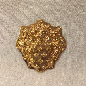 May include: A gold-colored metal button with a detailed design of a shield and a crest. The shield is decorated with a pattern of small diamonds.