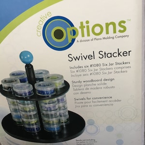 Puede incluir: Un apilador giratorio negro con seis tarros de plástico transparente llenos de cuentas de colores. El producto está etiquetado como "Swivel Stacker" e incluye texto de "Creative Options". El apilador tiene un diseño de tablero de madera resistente.